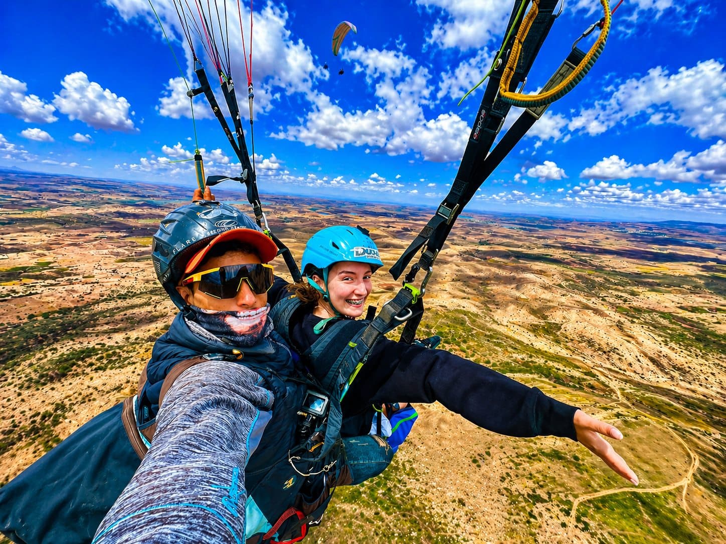 Atlas Paragliding tandem flight above wide mountain terrain under a bright blue sky.